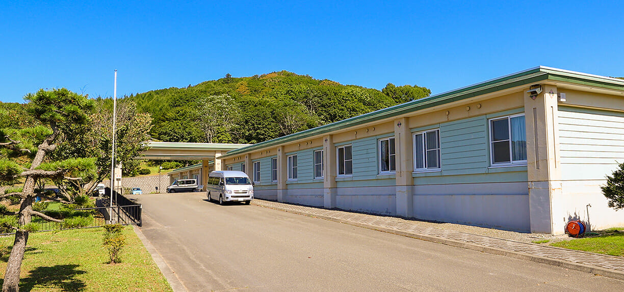 緑に囲まれた介護施設の建物外観。駐車場と車が見える。