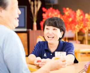 職員と入居者が笑顔で会話・交流している様子。温かみのある共用空間での日常風景。